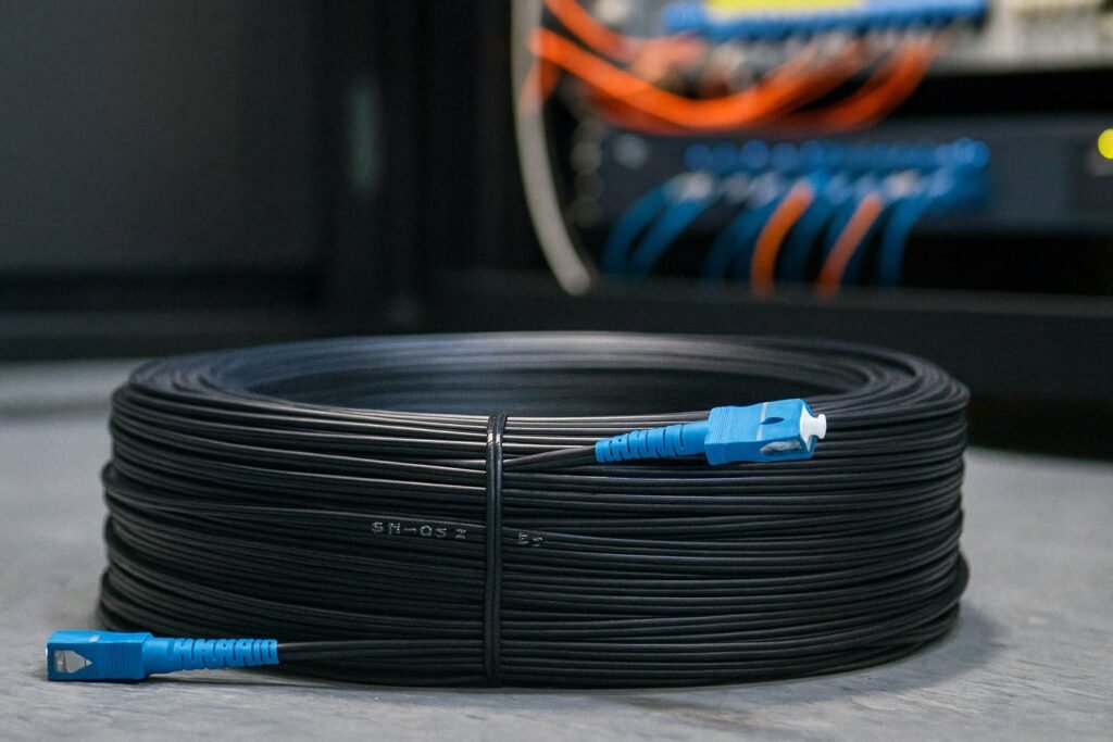 Neatly wound black pre-terminated fiber optic cable coil with blue SC connector in the foreground and a patch panel rack in the blurred background.